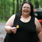 Fat white woman in a black dress with long black hair in a forest, with trees and a car behind her, holding a small yellow leaf and smiling.