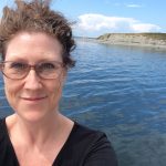 White woman with glasses and her hair in a messy bun in front of water and a distant spit of land.