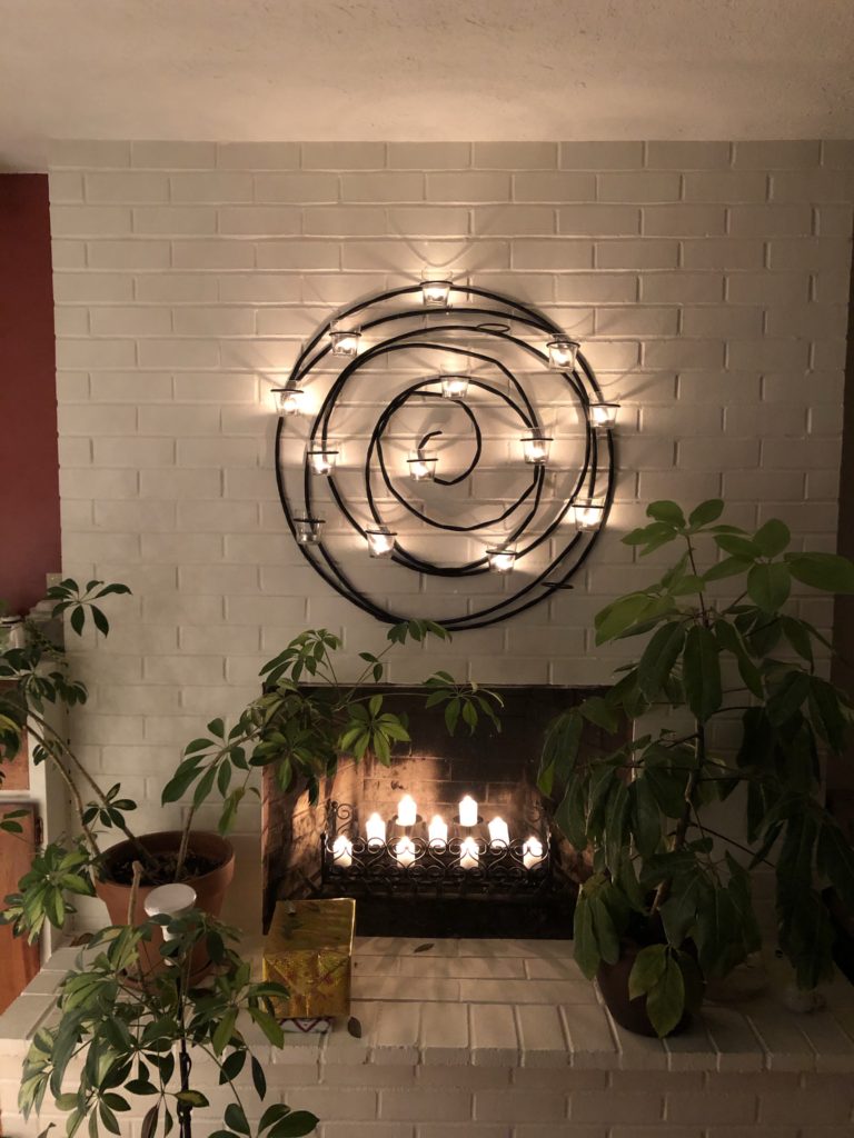 white brick fireplace with a black metal spiral lit with candles, a group of candles in the fireplace, and two big plants flanking the fireplace